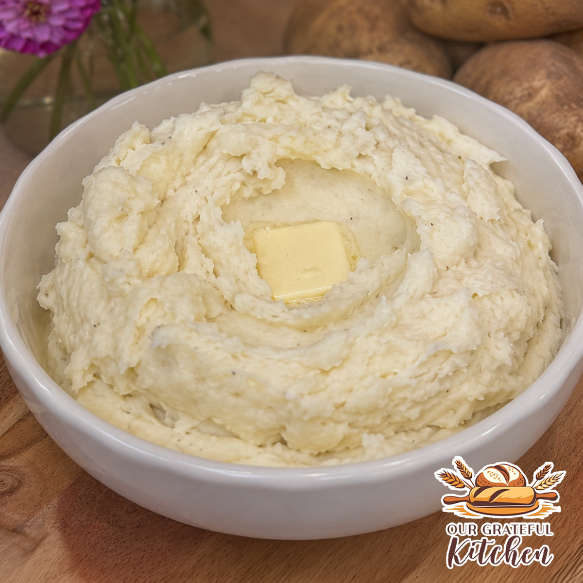 Creamy homemade mashed potatoes in a white serving bowl topped with melting butter on a wooden board with fresh flowers in the background.