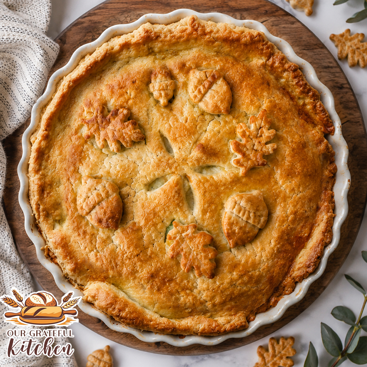Homemade chicken pot pie with a golden flaky crust decorated with acorn and oak leaf pie crust cutouts, baked in a white pie dish on a round wooden board over a white marble countertop.